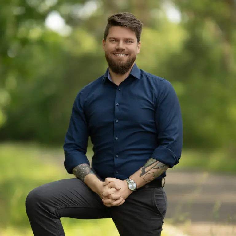 Business Portrait eines Mannes mit Bart und Tattoos im blauen Hemd, sitzend im Freien vor unscharfem Naturhintergrund, fotografiert von Ulrich Roth