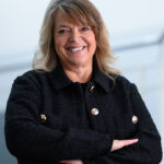 Businessporträt einer lächelnden Frau mit verschränkten Armen vor hellem Hintergrund.