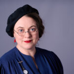 Professioneller Corporate Headshot einer Frau mit Brille und rotem Lippenstift vor grauem Hintergrund