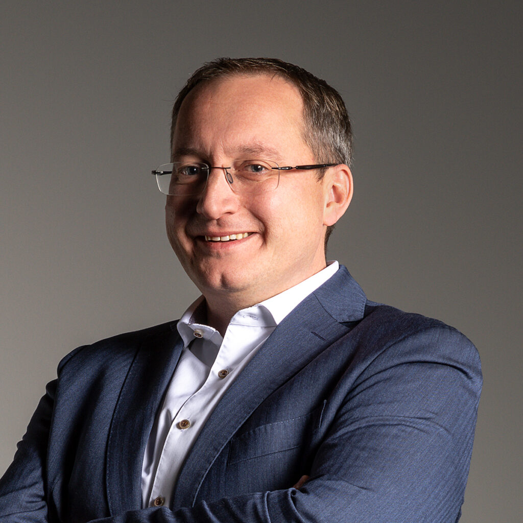 Business-Porträt eines Mannes mit Brille und blauem Sakko vor grauem Hintergrund.