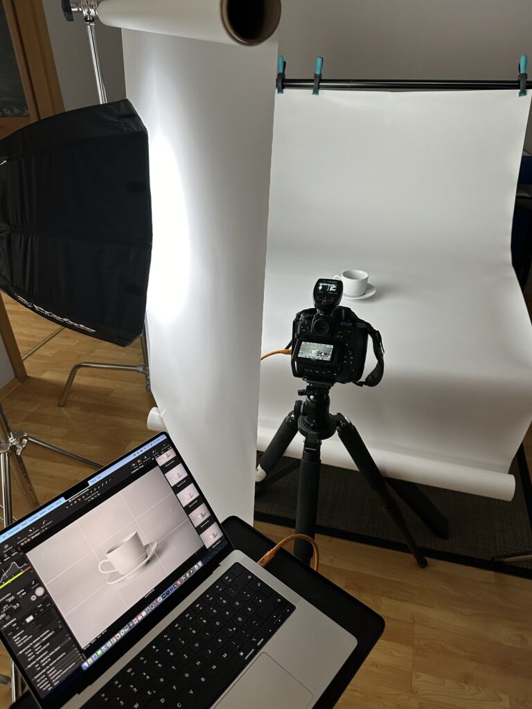 Kamera auf Stativ vor einer weißen Tasse auf Papierhintergrund, Laptop zeigt das Bild