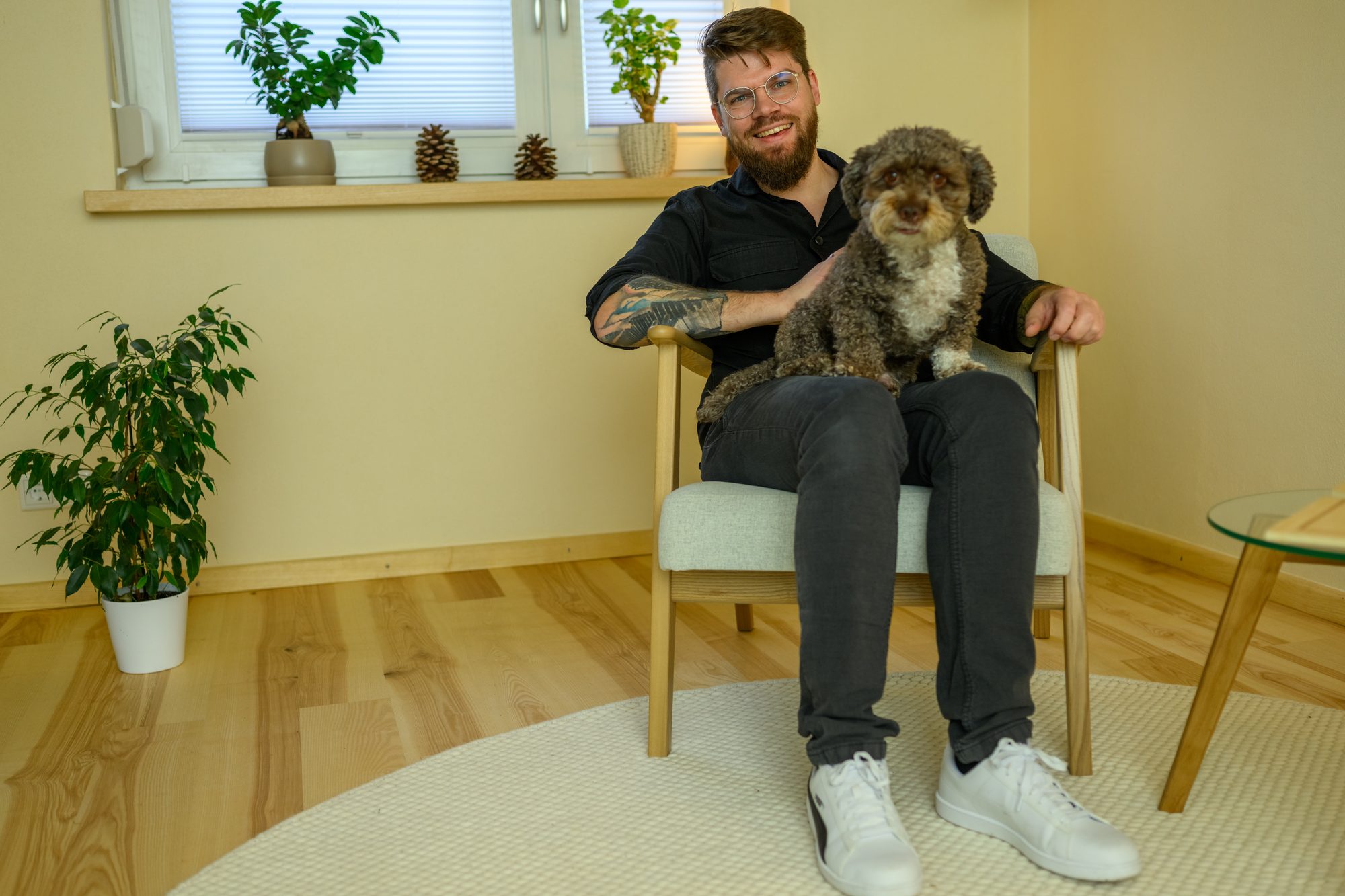 Ronny Mosebach sitzt lachend mit seinem lockigen braunen Hund auf dem Schoß im Sessel seiner Praxis